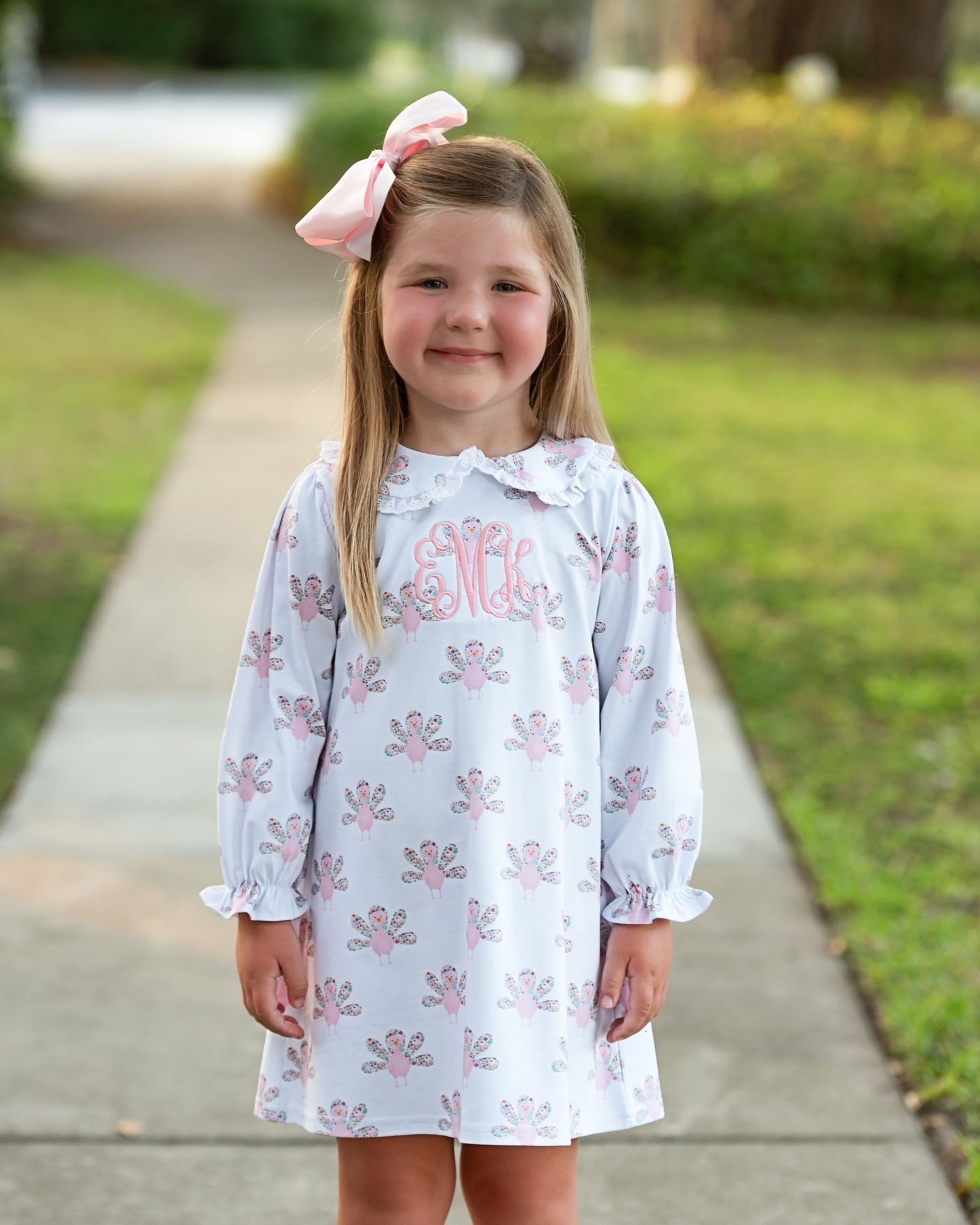 Young girl in a white dress with pink Turkey patterns and a large pink bow outdoors.