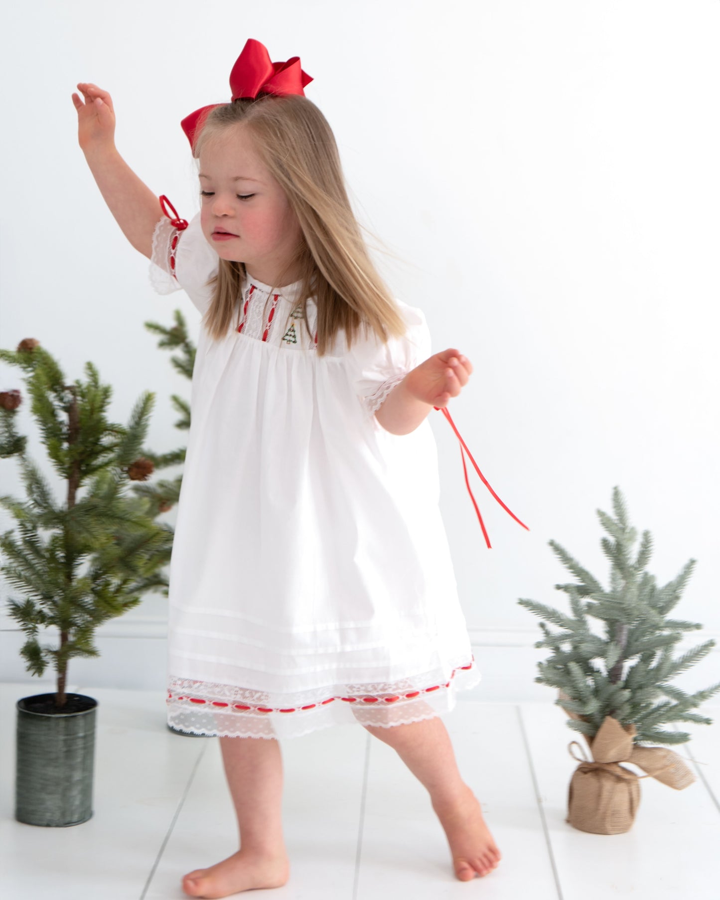 Young girl in a white dress with red accents standing between two small Christmas trees.