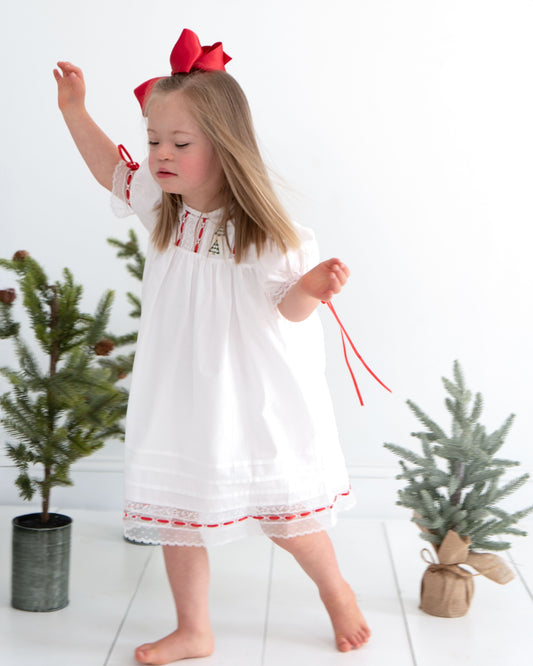 Young girl in a white dress with red accents standing between two small Christmas trees.