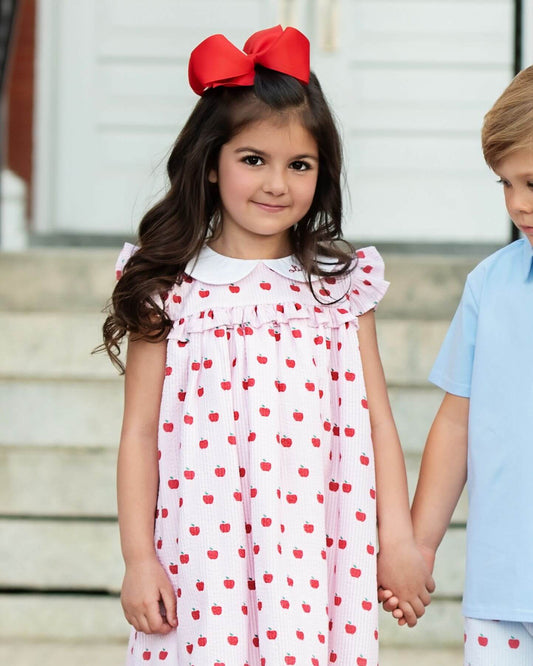 Young girl in a white dress with red cherries and a red bow, standing outdoors.