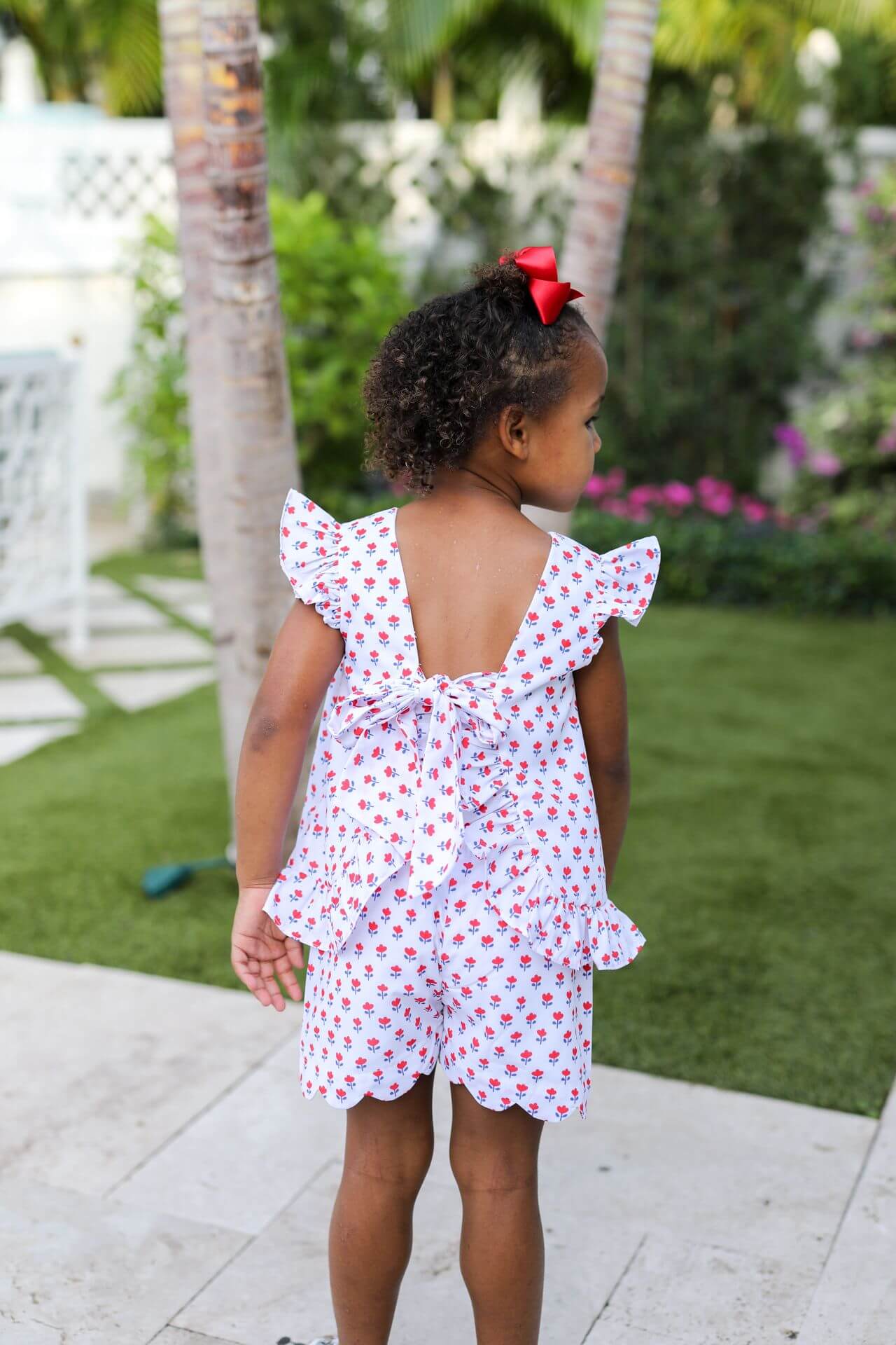 Young girl in a white dress with red patterns standing outdoors on a patio.