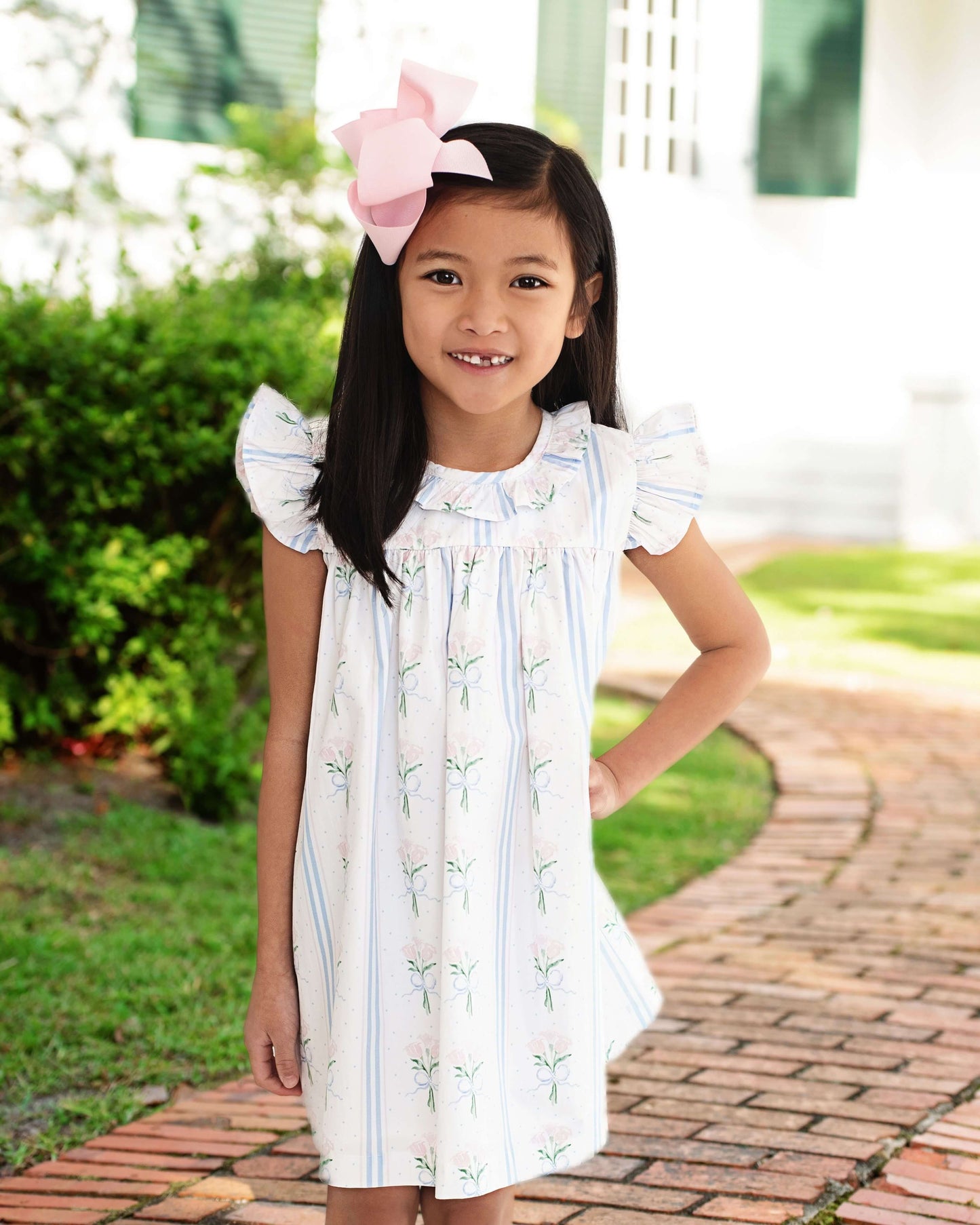 Young girl in a white dress with ruffled sleeves standing outdoors on a brick path.