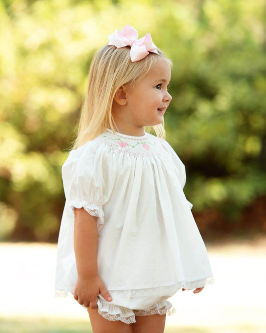 Young girl in a white outfit with smocked hearts, a pink bow in her hair, standing outdoors.
