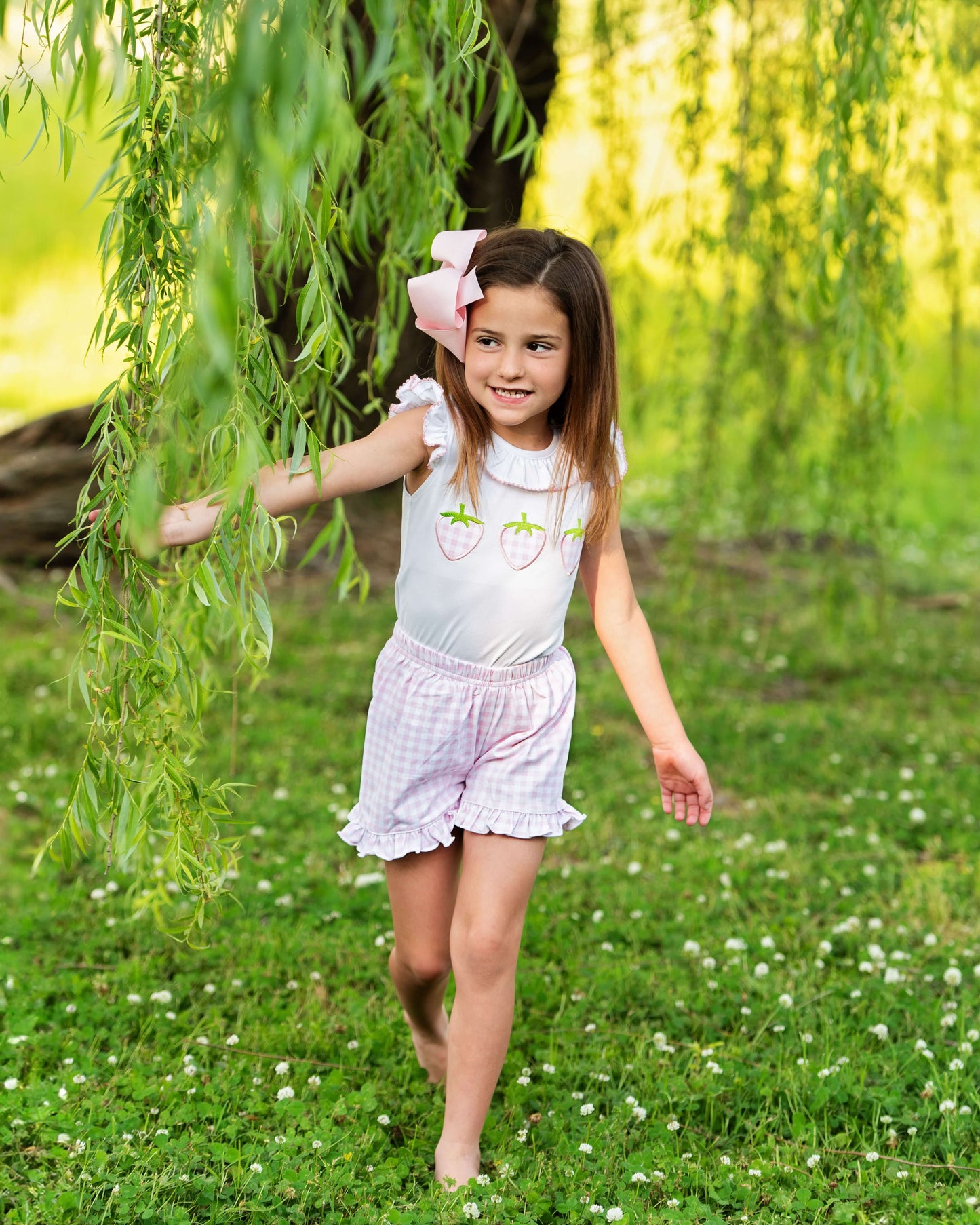 Young girl in a white top and pink shorts standing under a willow tree in a grassy field.