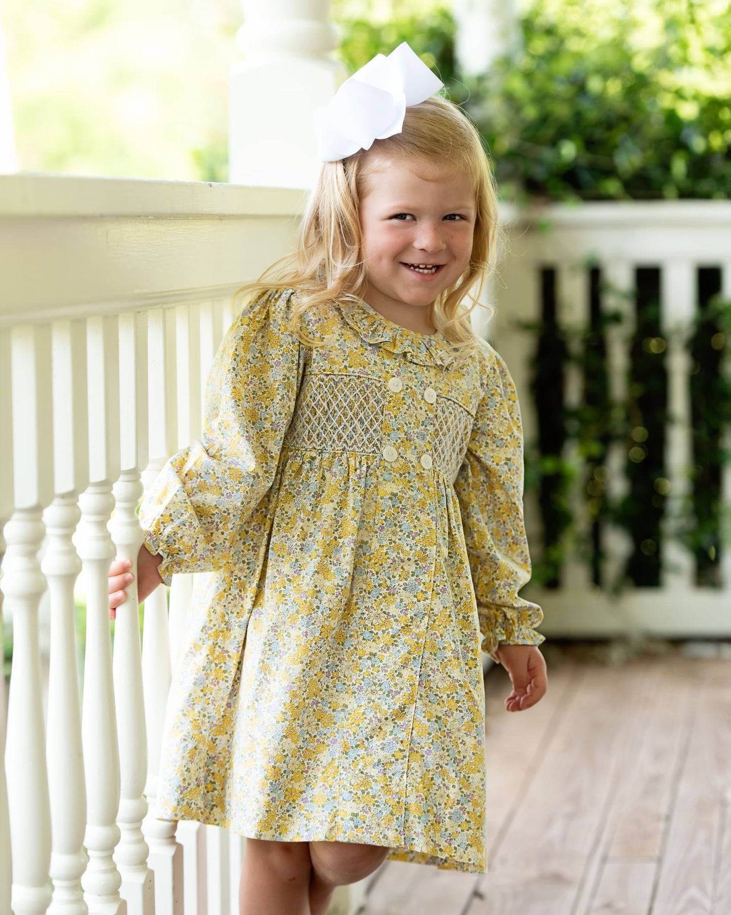 Young girl in a yellow floral dress standing on a porch.