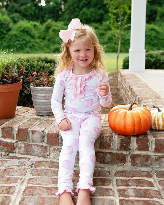 Young girl in pink and white outfit with a large bow, sitting on brick steps with pumpkins and plants around.