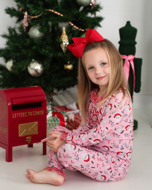 Young girl in pink pajamas with a red bow sitting in front of a Christmas tree and red mailbox.