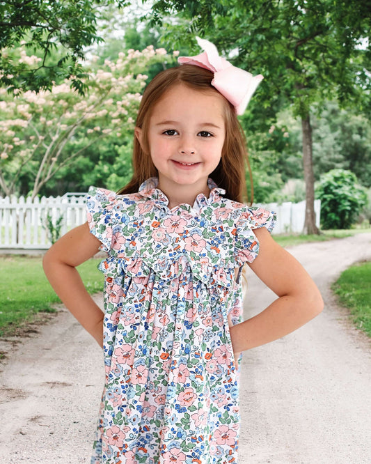 Young girl posing in a floral dress, wearing a pink bow in her hair, standing outside