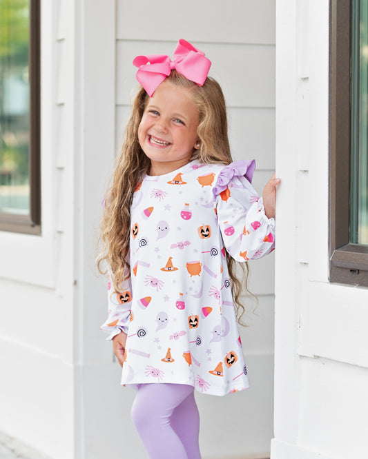 Young girl smiling wearing a Halloween-themed legging set with a pink bow in her hair, standing in front of a white door.