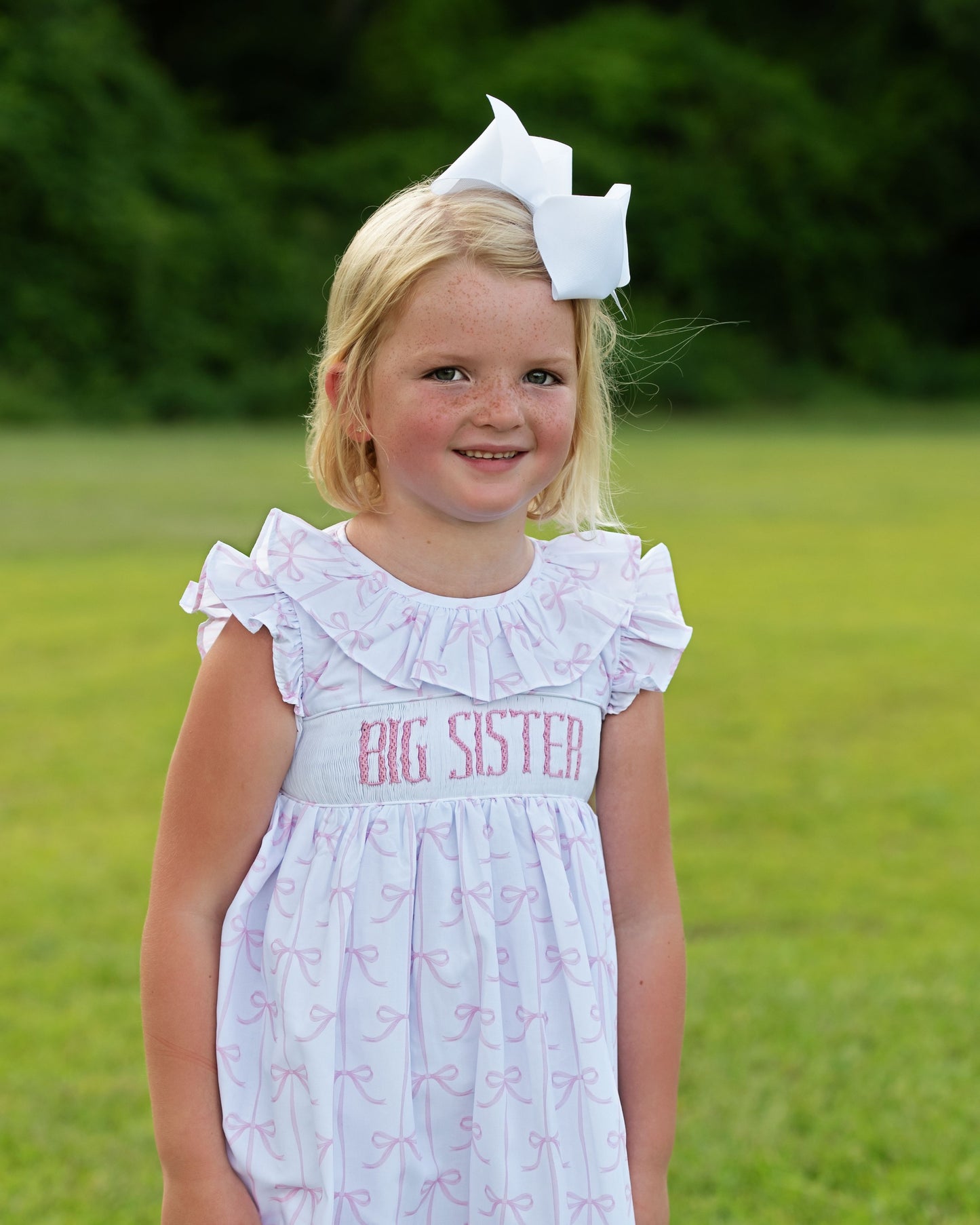 Young girl wearing a 'Big Sister' dress with a large white bow in her hair, standing in a grassy field.
