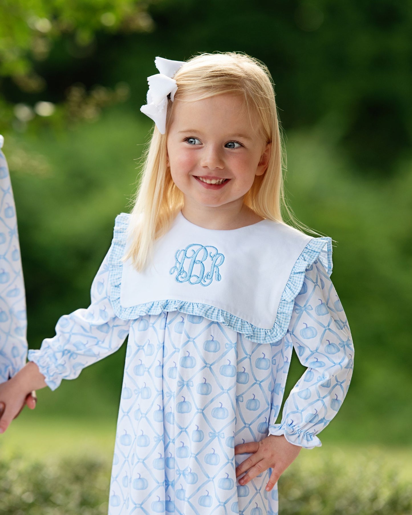 Young girl wearing a blue and white dress with a monogram, standing outdoors.