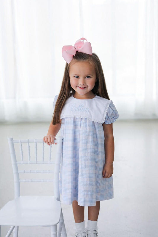 Young girl wearing a blue checkered dress with a white collar, standing in a room with a white chair and light background.