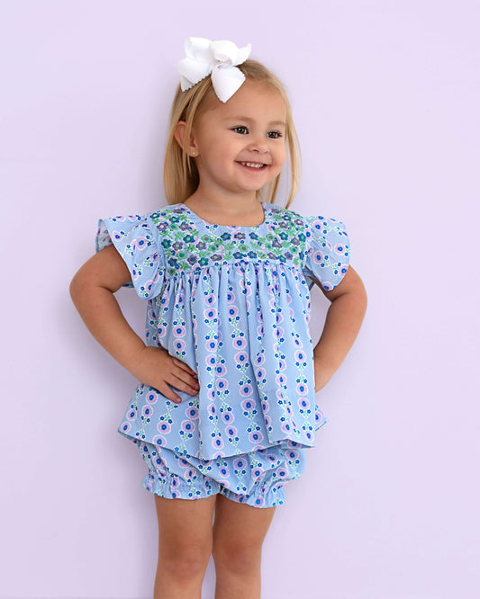 Young girl wearing a blue floral outfit with a white bow against a light purple background