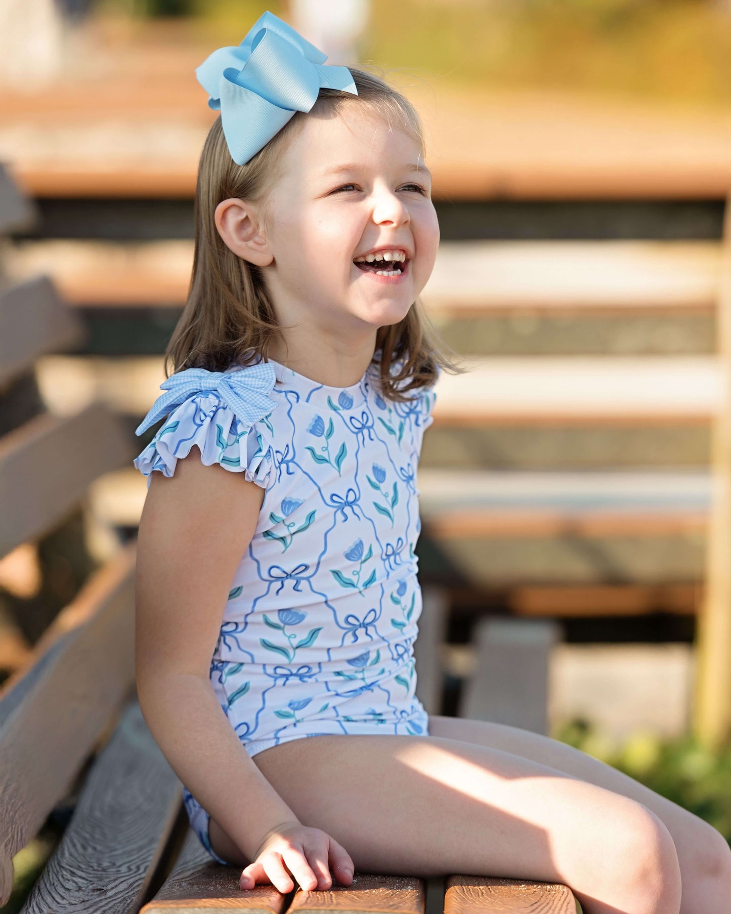 Young girl wearing a blue floral swimsuit with a large bow in her hair, sitting on wooden steps.