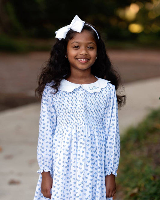 Young girl wearing a blue patterned dress with a white bow in her hair, standing outdoors.