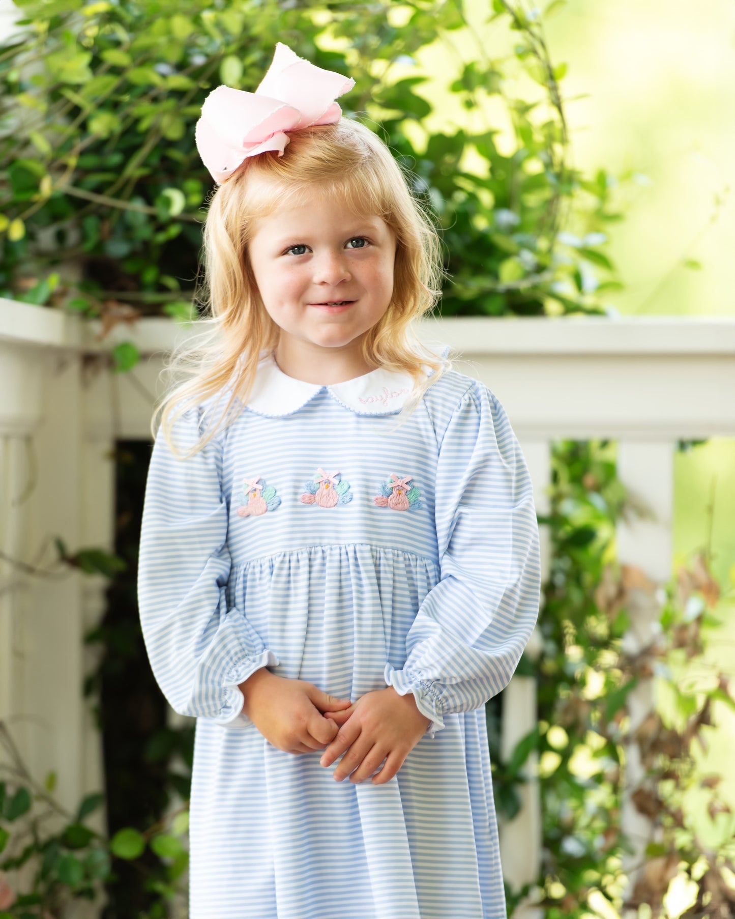 Young girl wearing a blue striped dress with embroidered turkeys and a pink bow in her hair, standing outdoors.