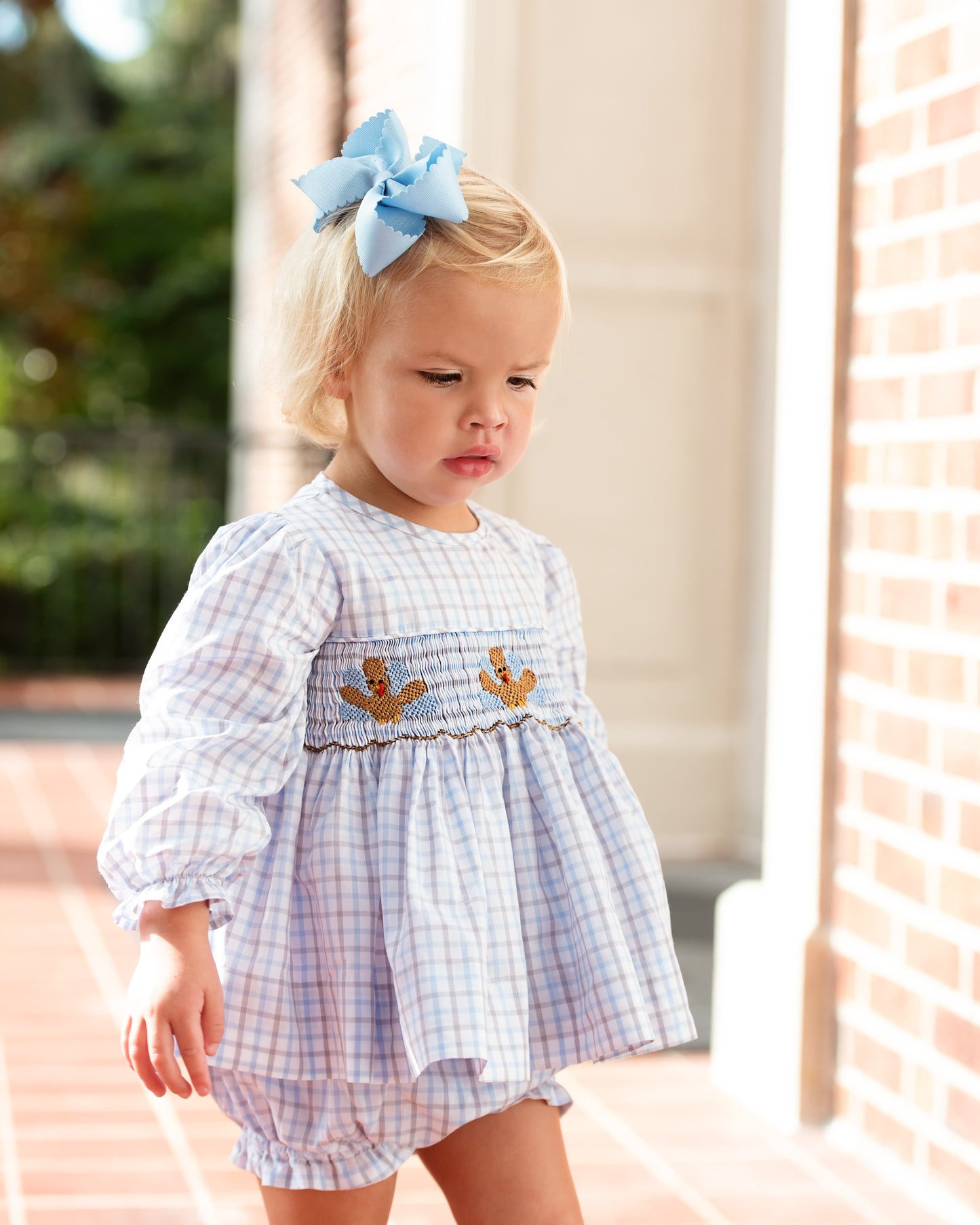 Young girl wearing a checkered diaper set with smocked turkeys outdoors.