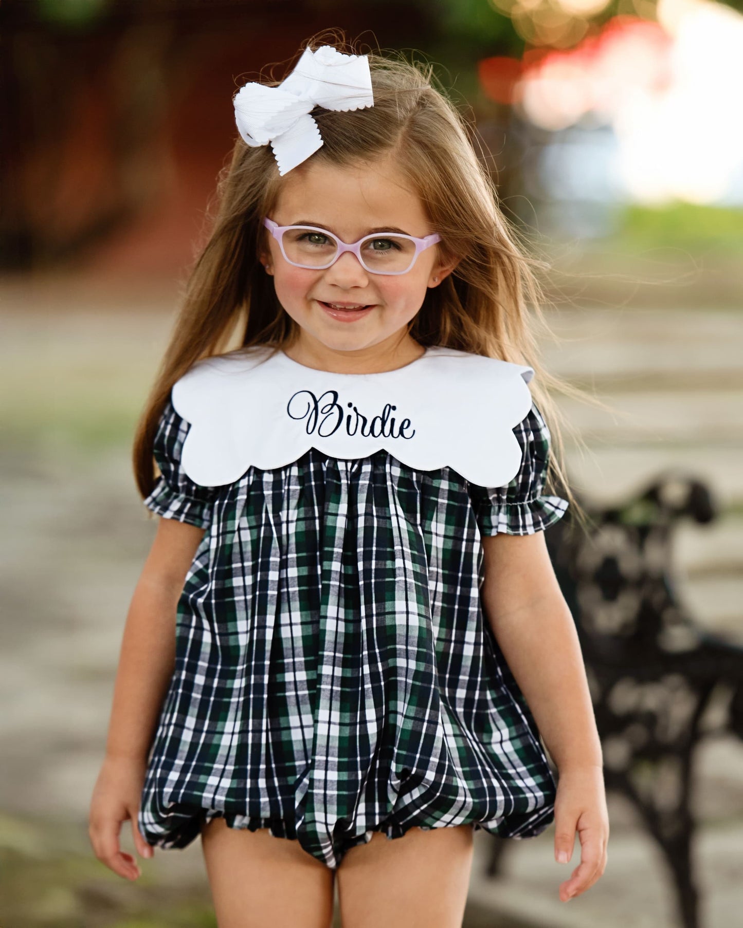 Young girl wearing a checkered romper with 'Birdie' on it, standing outdoors.