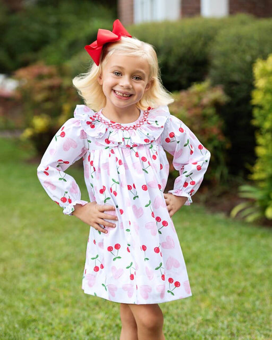Young girl wearing a cherry-patterned dress with a red bow outdoors.