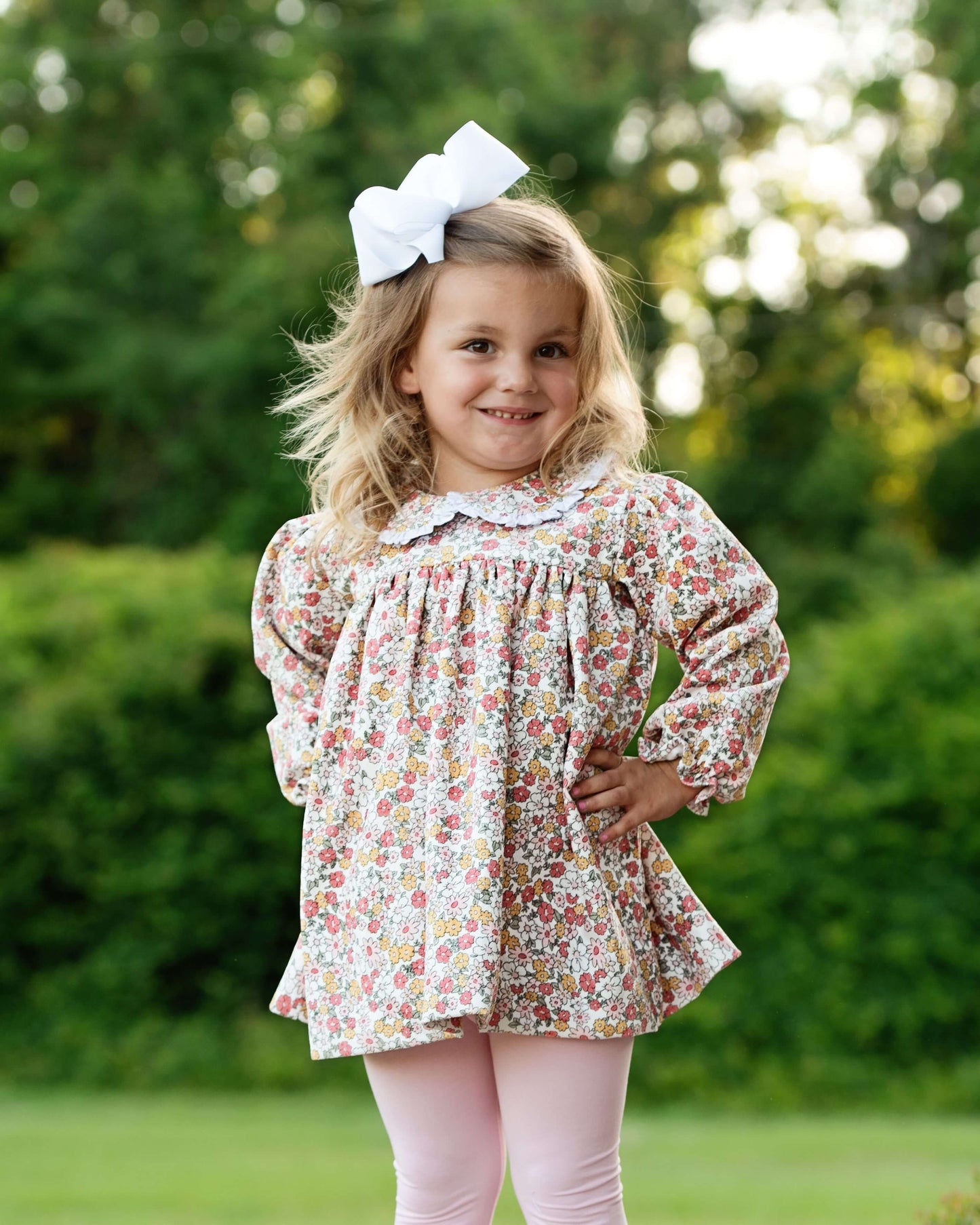 Young girl wearing a floral blouse and pink pants with a white bow in her hair, standing outdoors.