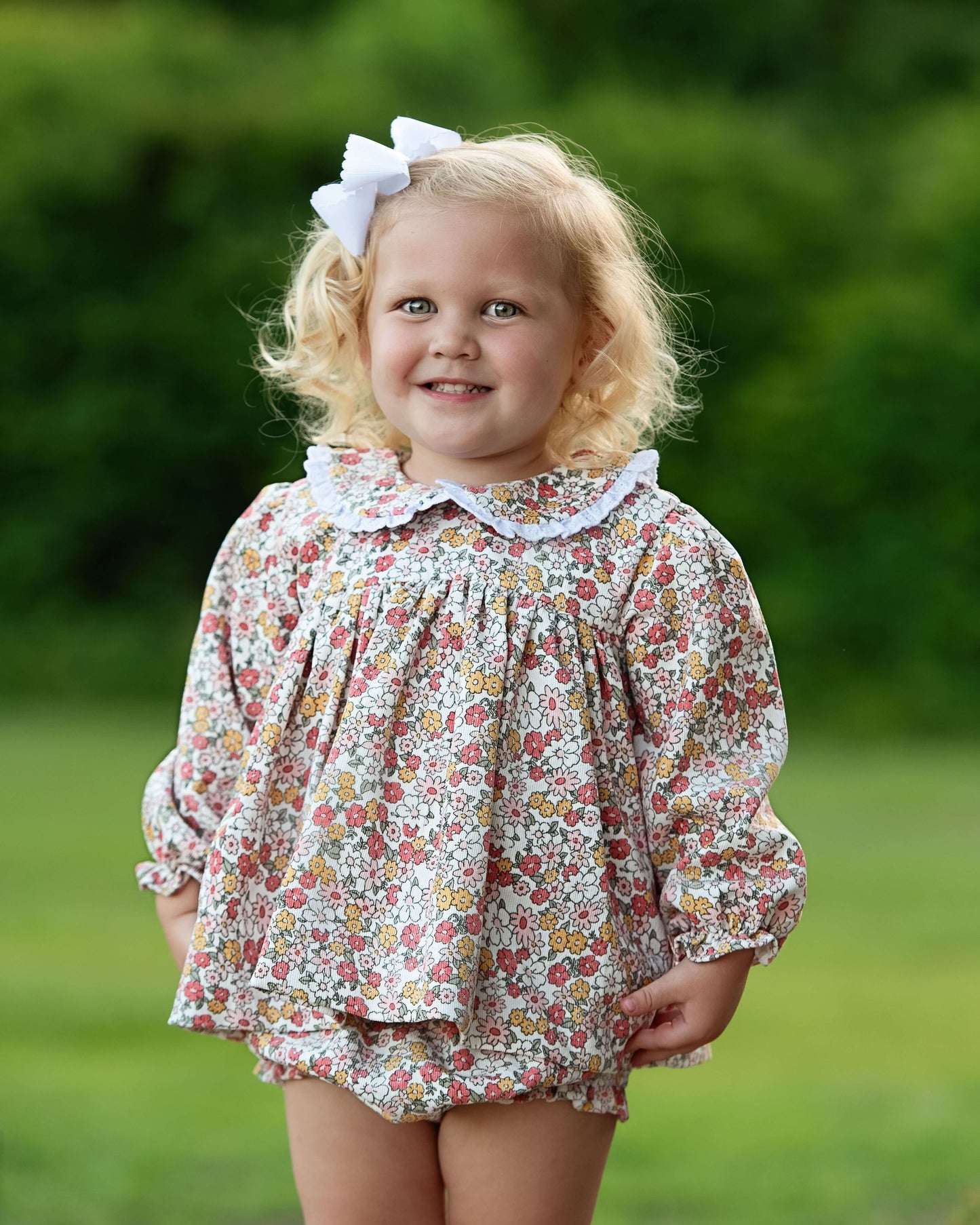 Young girl wearing a floral blouse and shorts with a white bow outdoors.
