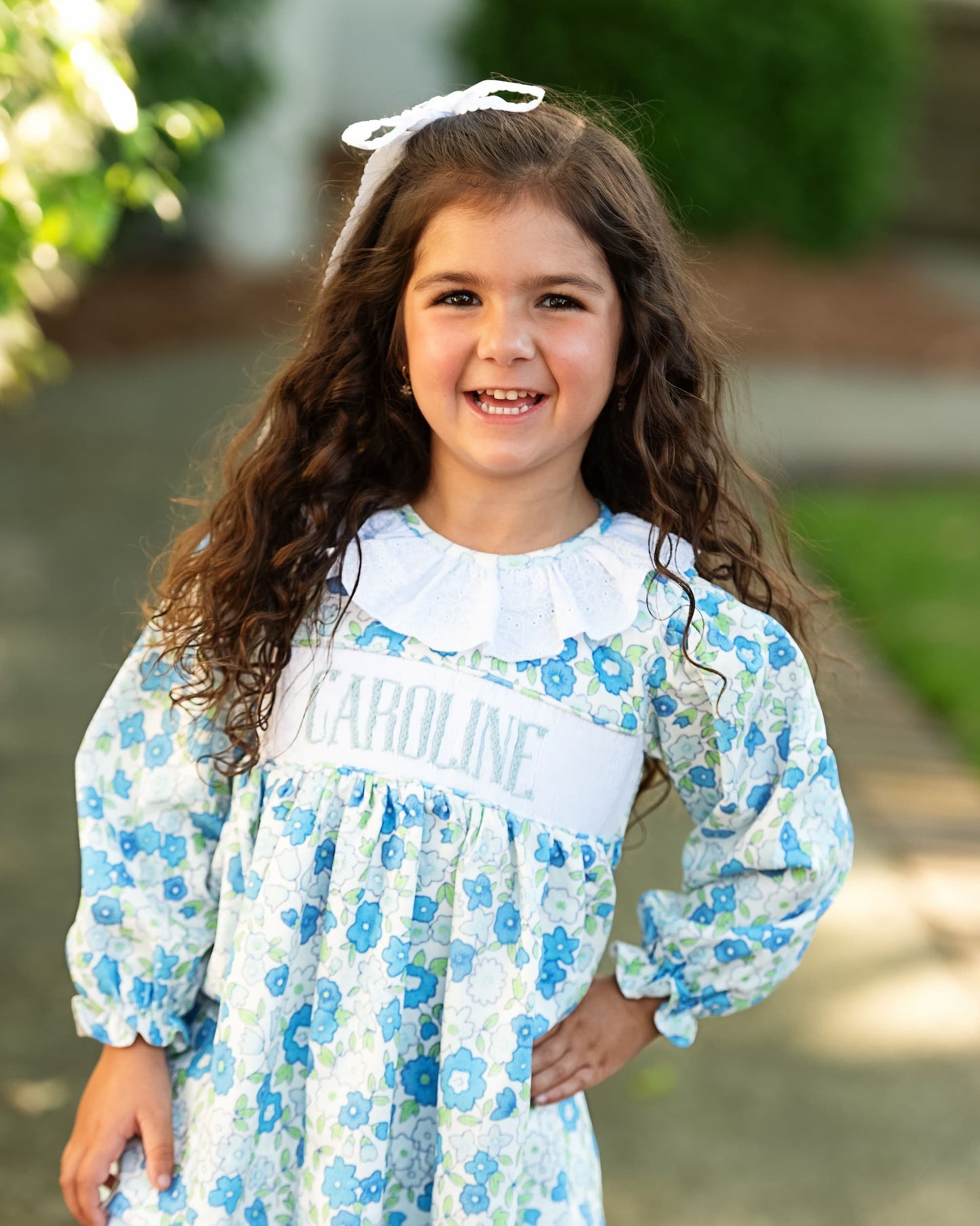 Young girl wearing a floral dress with 'Caroline' embroidered on it, standing outdoors.