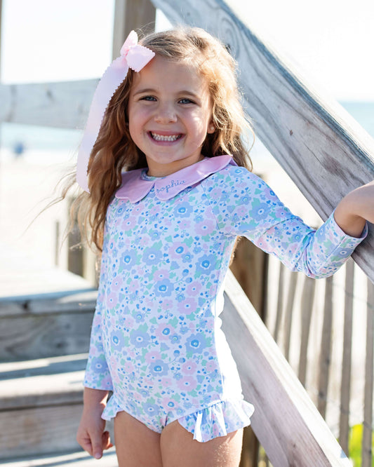 Young girl wearing a floral dress with a collar, standing on a wooden deck.