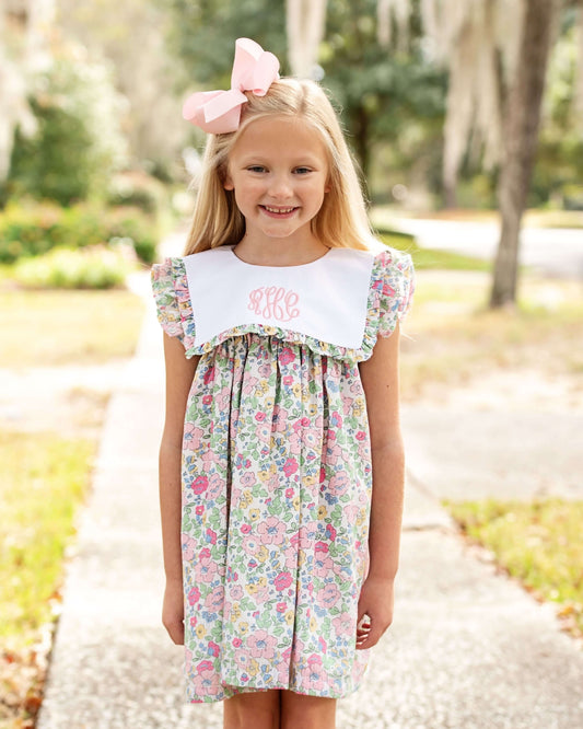 Young girl wearing a floral dress with a white collar and pink bow in her hair, standing outdoors.
