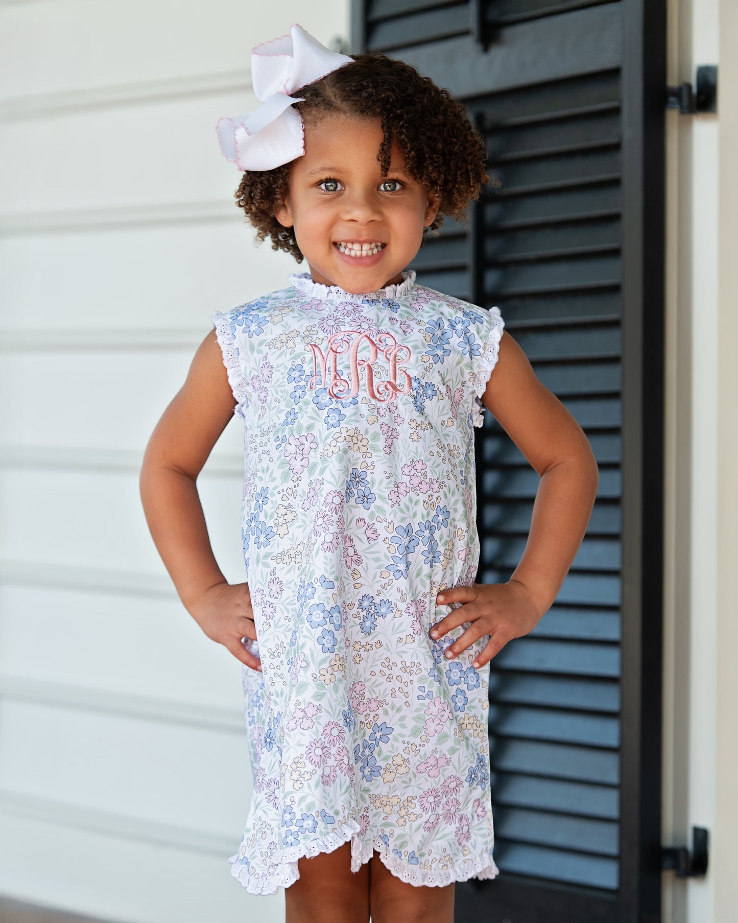 Young girl wearing a floral dress with monogram, standing in front of a white wall and black shutters.