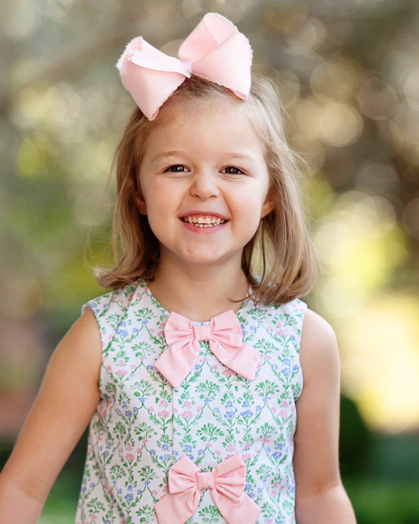 Young girl wearing a floral dress with pink bows against a blurred natural background