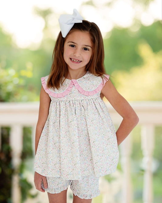 Young girl wearing a floral dress with pink trim and a white bow in her hair, standing outdoors.