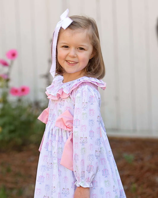 Young girl wearing a floral dress with ruffles and a pink bow, standing outdoors with flowers in the background.