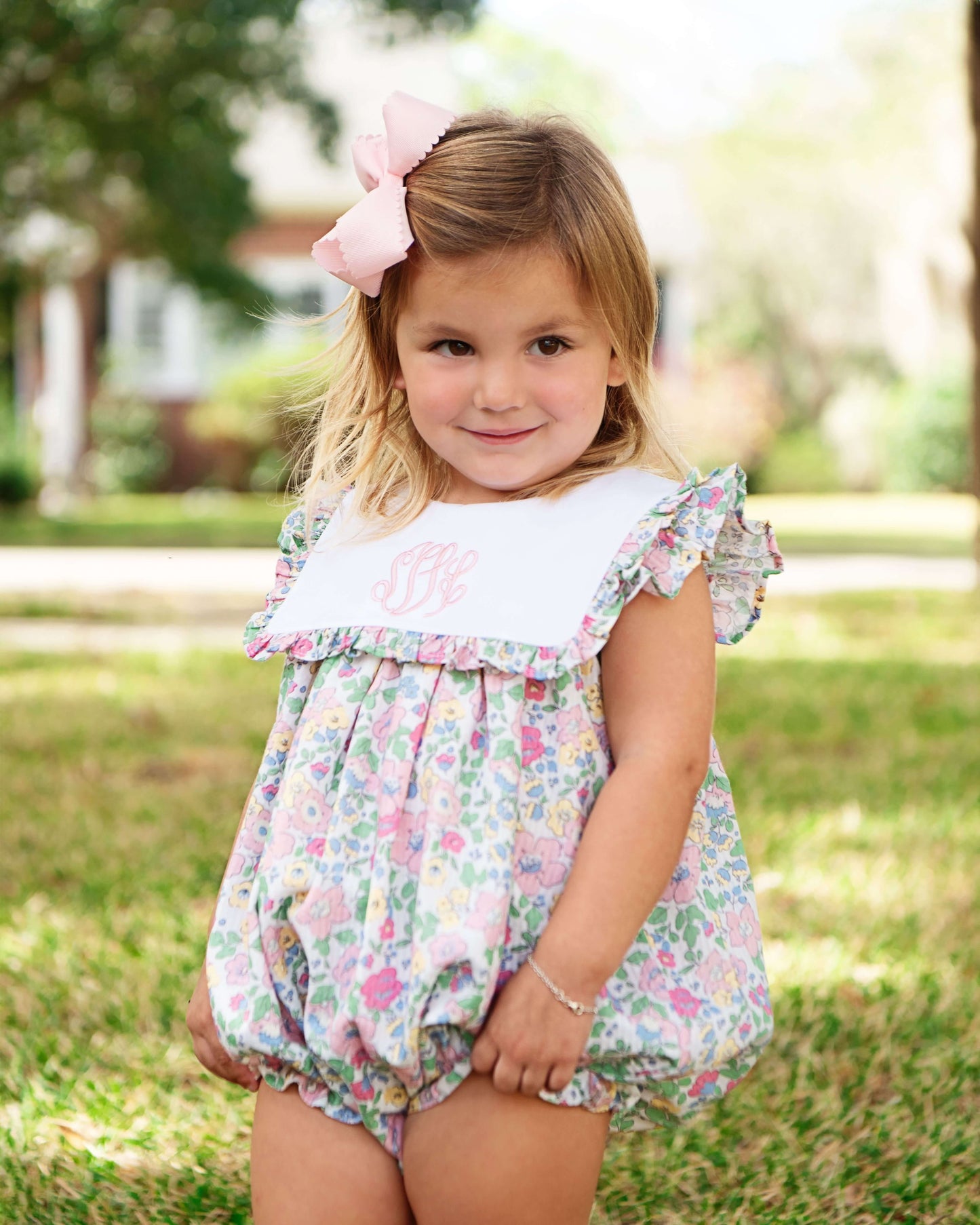 Young girl wearing a floral romper with a pink bow in her hair, standing outdoors.