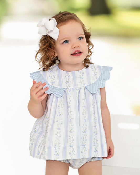Young girl wearing a light blue and white dress with ruffled details, holding a small object.