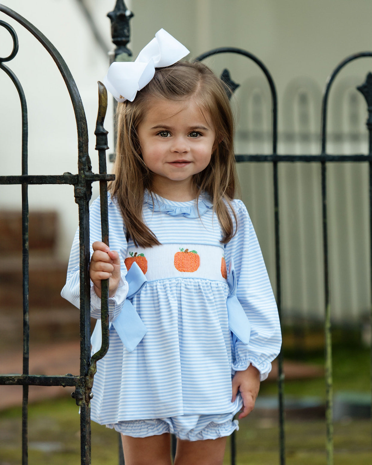 Young girl wearing a light blue diaper set with pumpkin design, standing in front of a black metal gate.