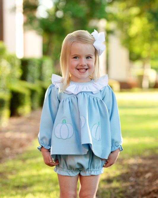 Young girl wearing a light blue diaper set with pumpkin designs and green shorts outdoors.