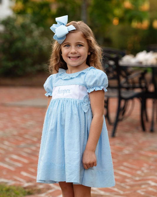 Young girl wearing a light blue dress with a large bow in her hair, standing outdoors on a brick path.