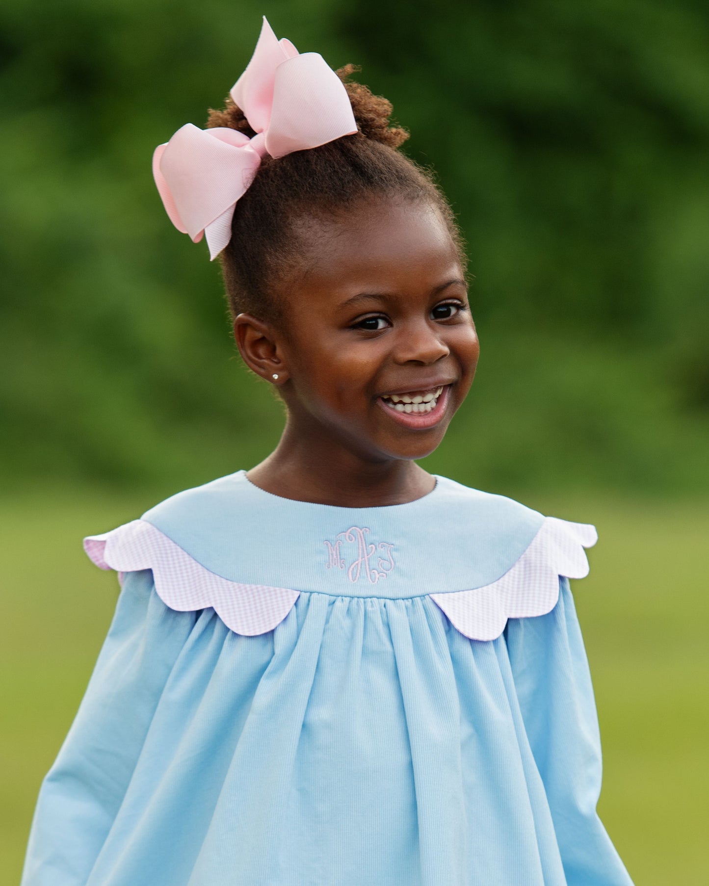 Young girl wearing a light blue dress with a white collar and pink bow in her hair, standing outdoors.