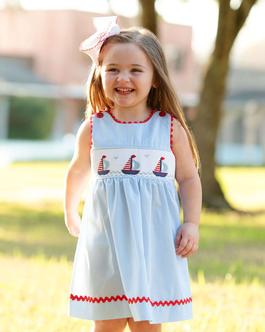 Young girl wearing a light blue dress with red accents and sailboat patterns outdoors.