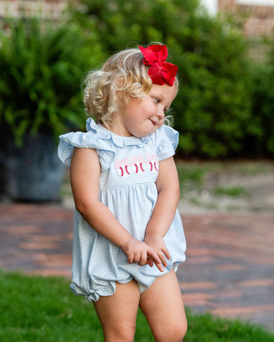 Young girl wearing a light blue dress with red lettering and a red bow outdoors.
