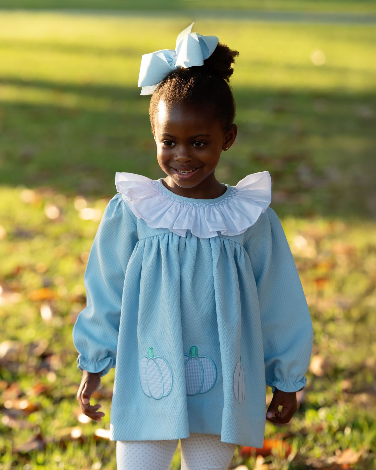 Young girl wearing a light blue legging set with white ruffled collar and pumpkin designs, standing outdoors on grass.