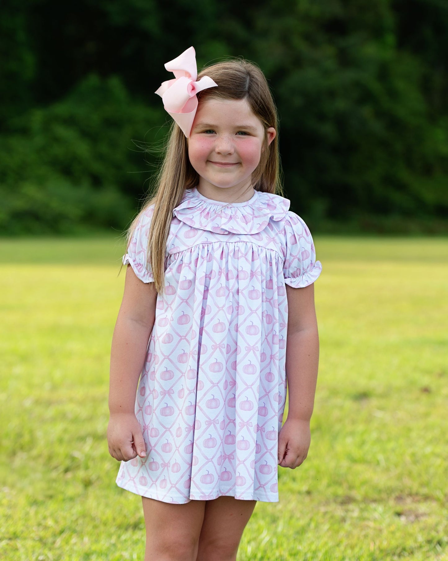 Young girl wearing a light purple dress with a floral pattern and a pink bow in her hair, standing in a grassy field.