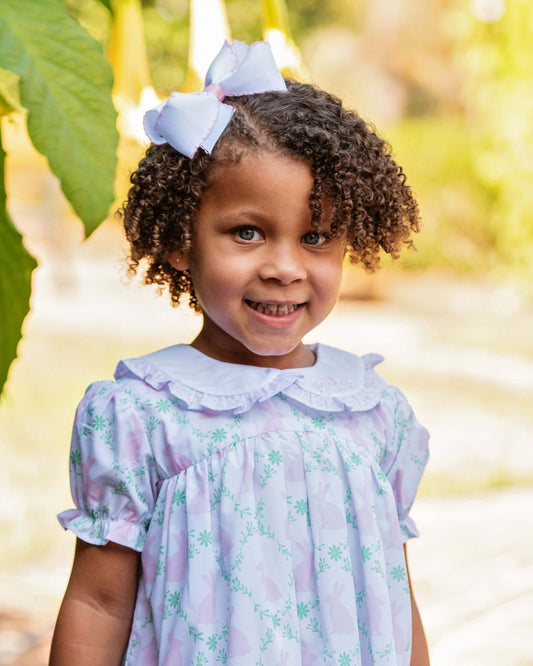 Young girl wearing a light purple dress with a floral pattern, standing outdoors with greenery in the background.