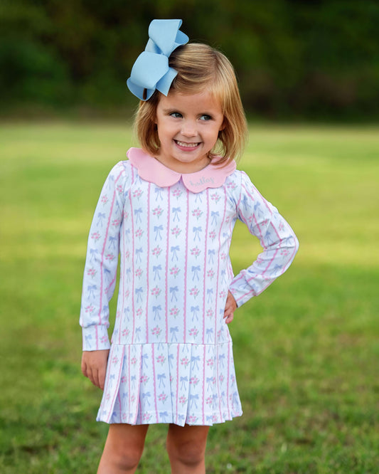 Young girl wearing a patterned dress with a pink collar and blue bow in a grassy outdoor setting
