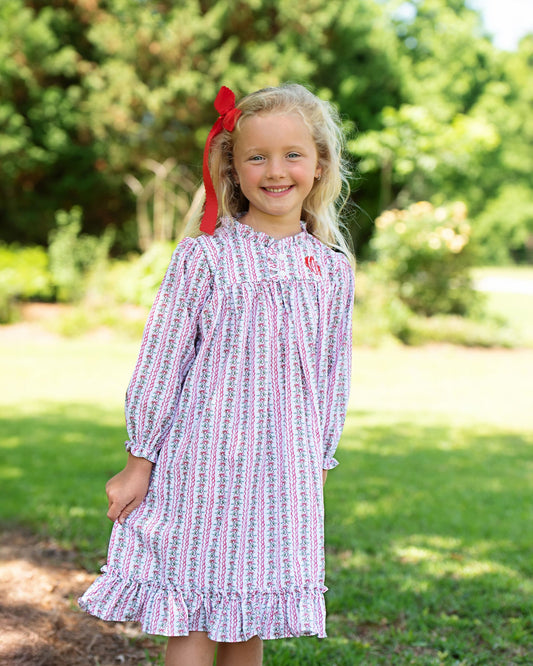 Young girl wearing a patterned dress with a red bow in her hair, standing outdoors.