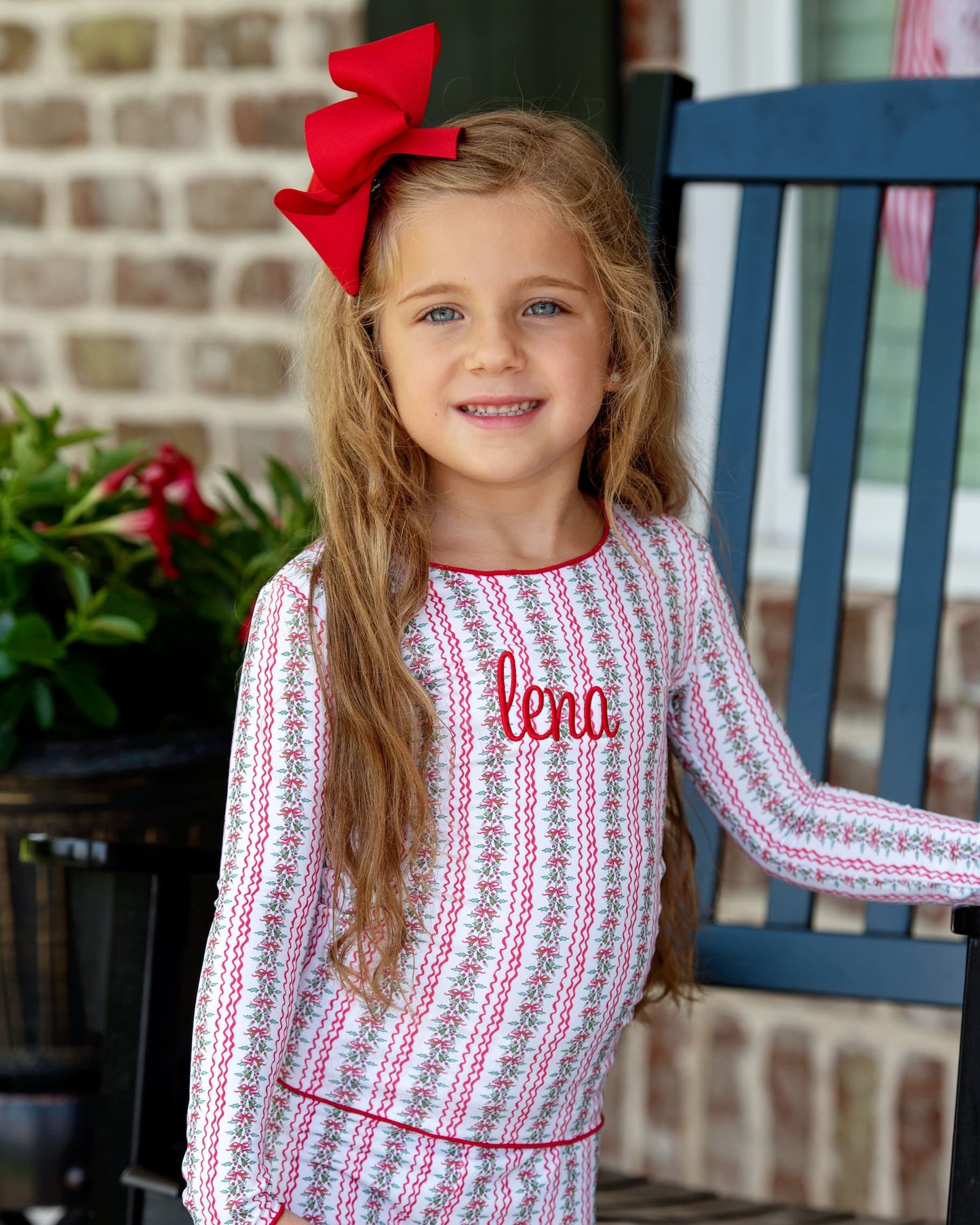 Young girl wearing a patterned dress with a red bow, standing on a porch.