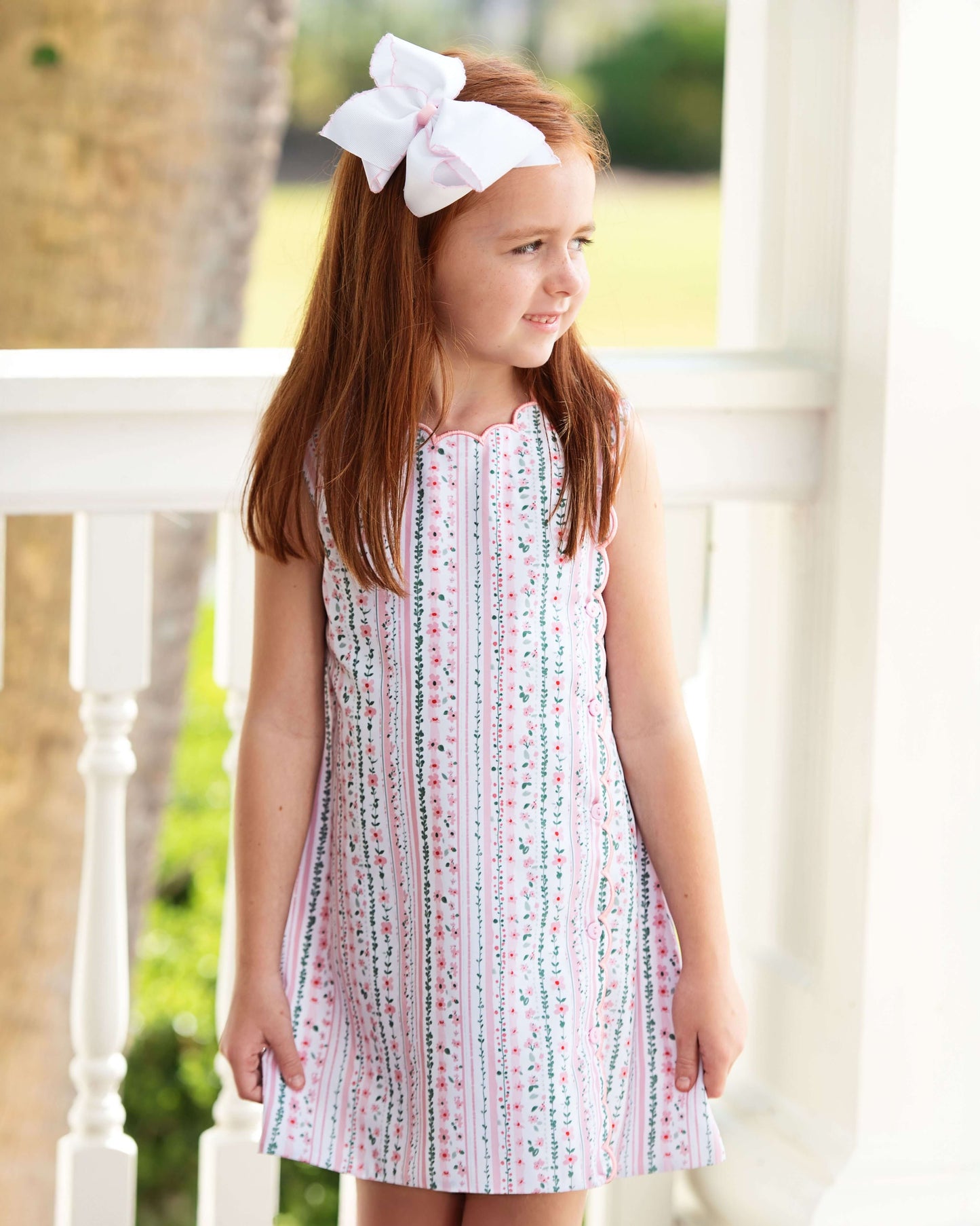 Young girl wearing a patterned dress with a white bow in her hair, standing on a porch.