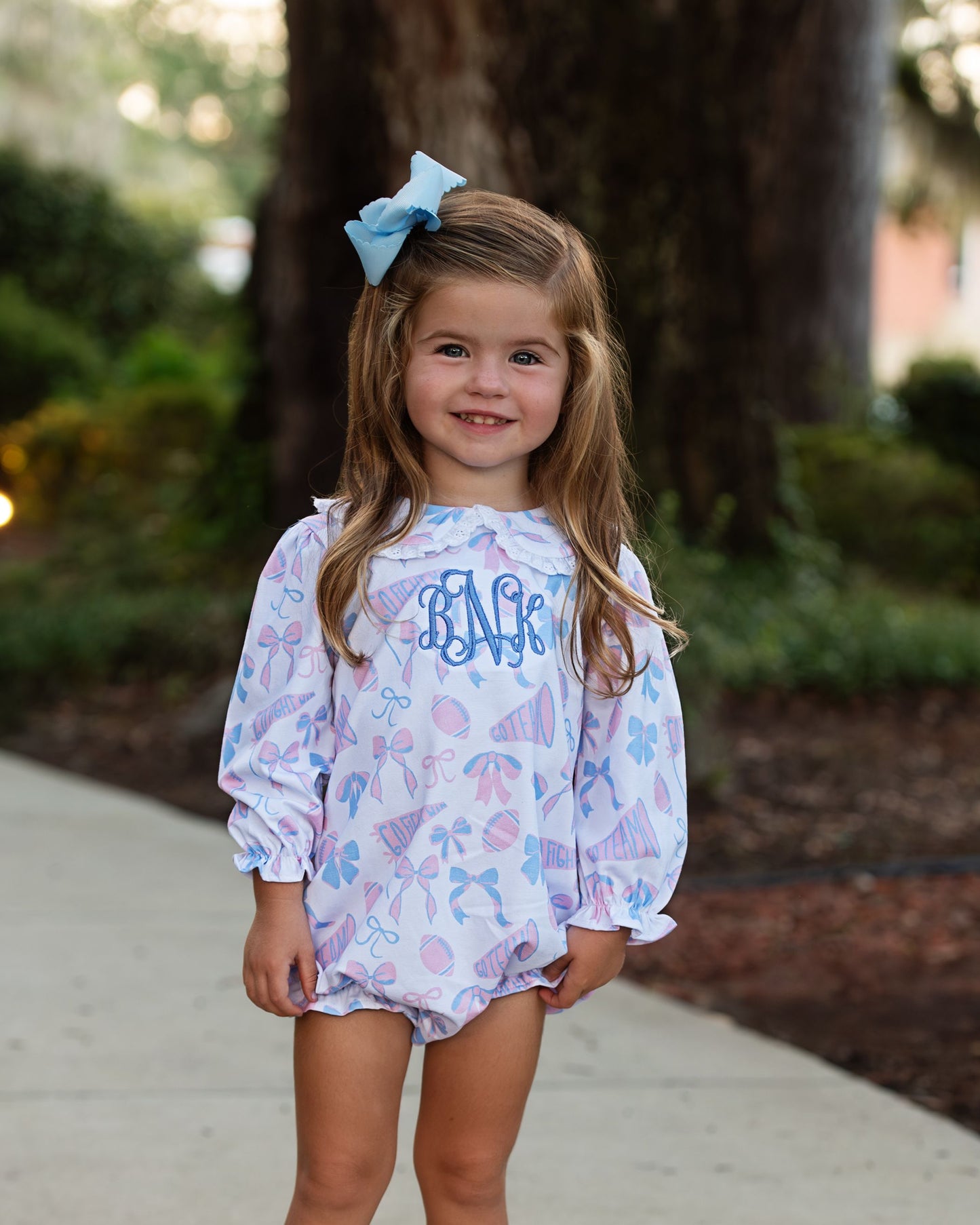 Young girl wearing a patterned outfit with 'Bella' monogram, standing outdoors.