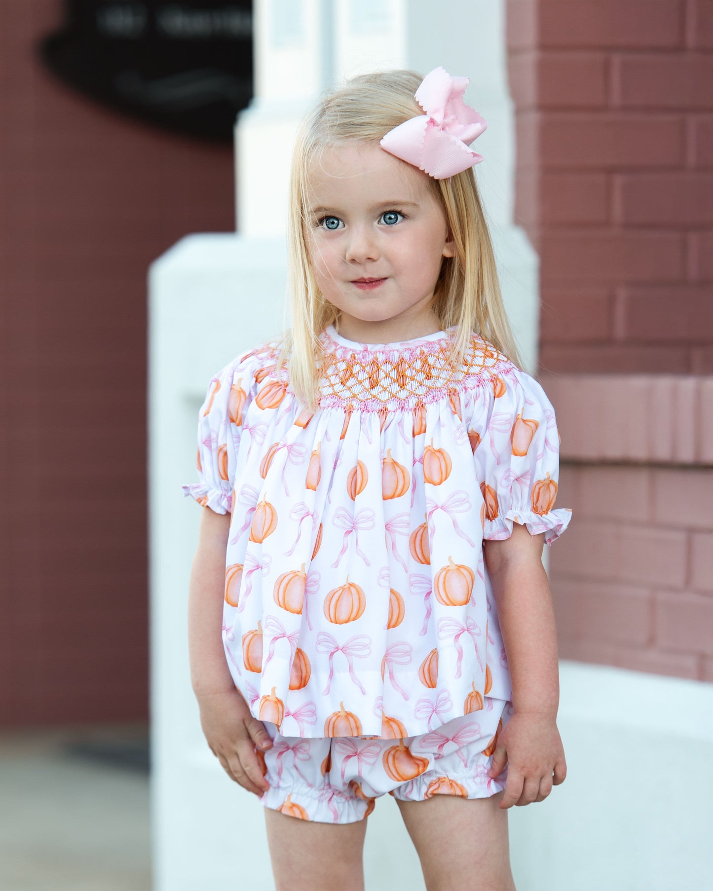 Young girl wearing a patterned outfit with a pink bow and pumpkin in her hair, standing outdoors 1