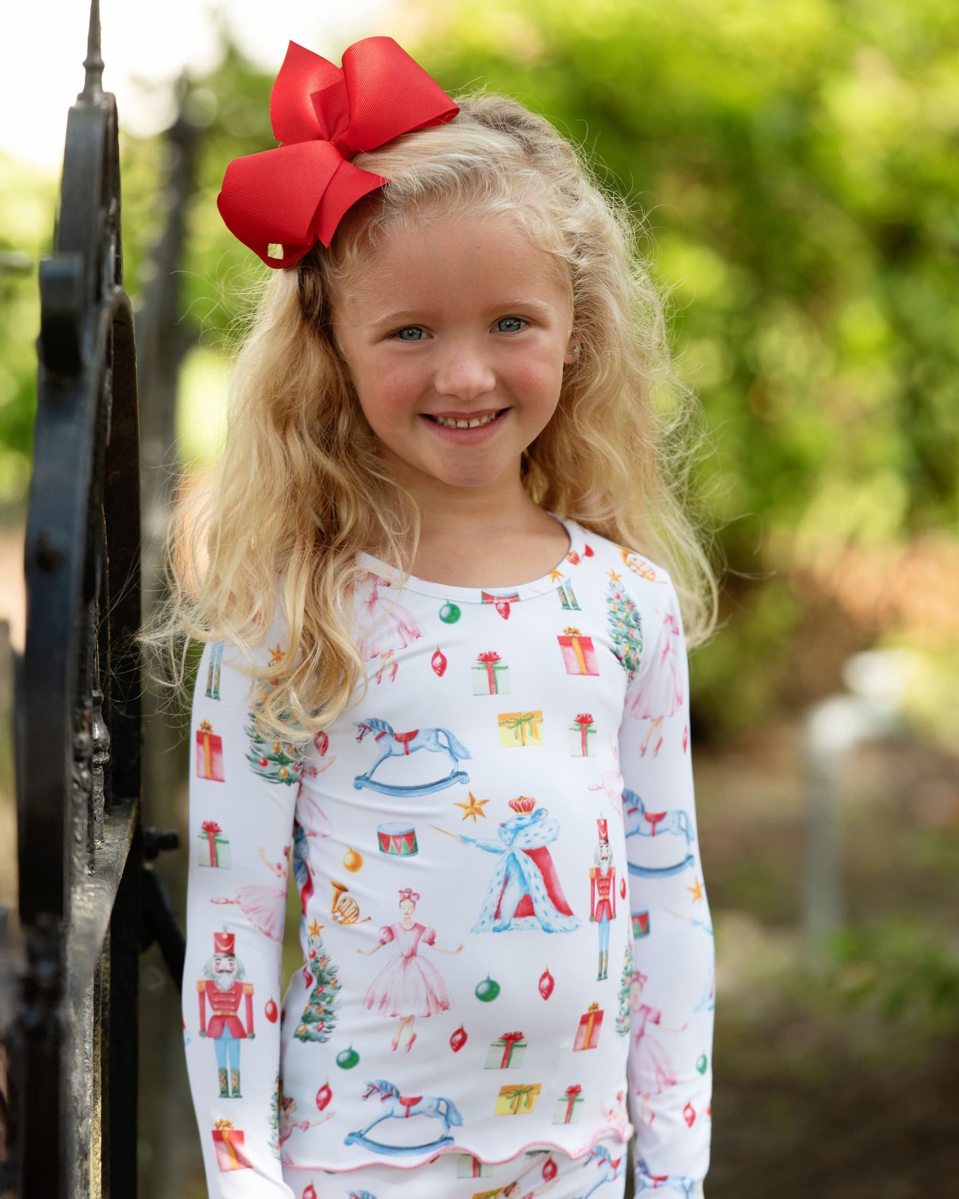 Young girl wearing a patterned shirt with a red bow in her hair, standing outdoors.